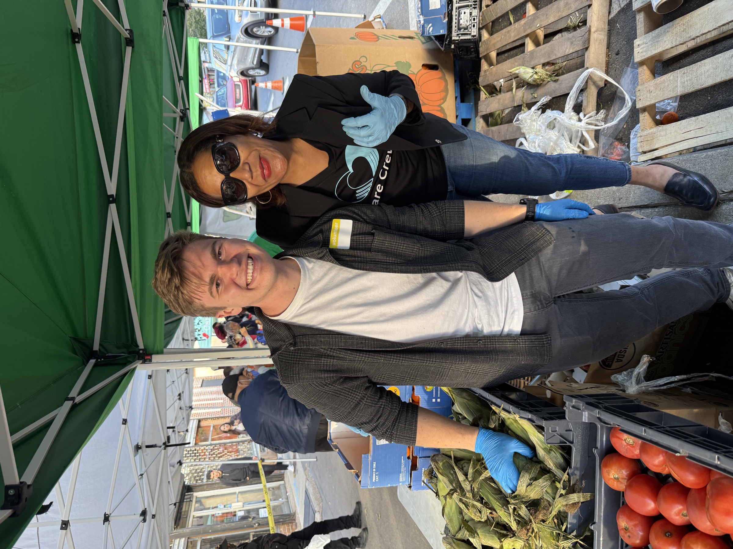 David volunteering at a street produce market, blue gloves on, crates of corn and tomatoes